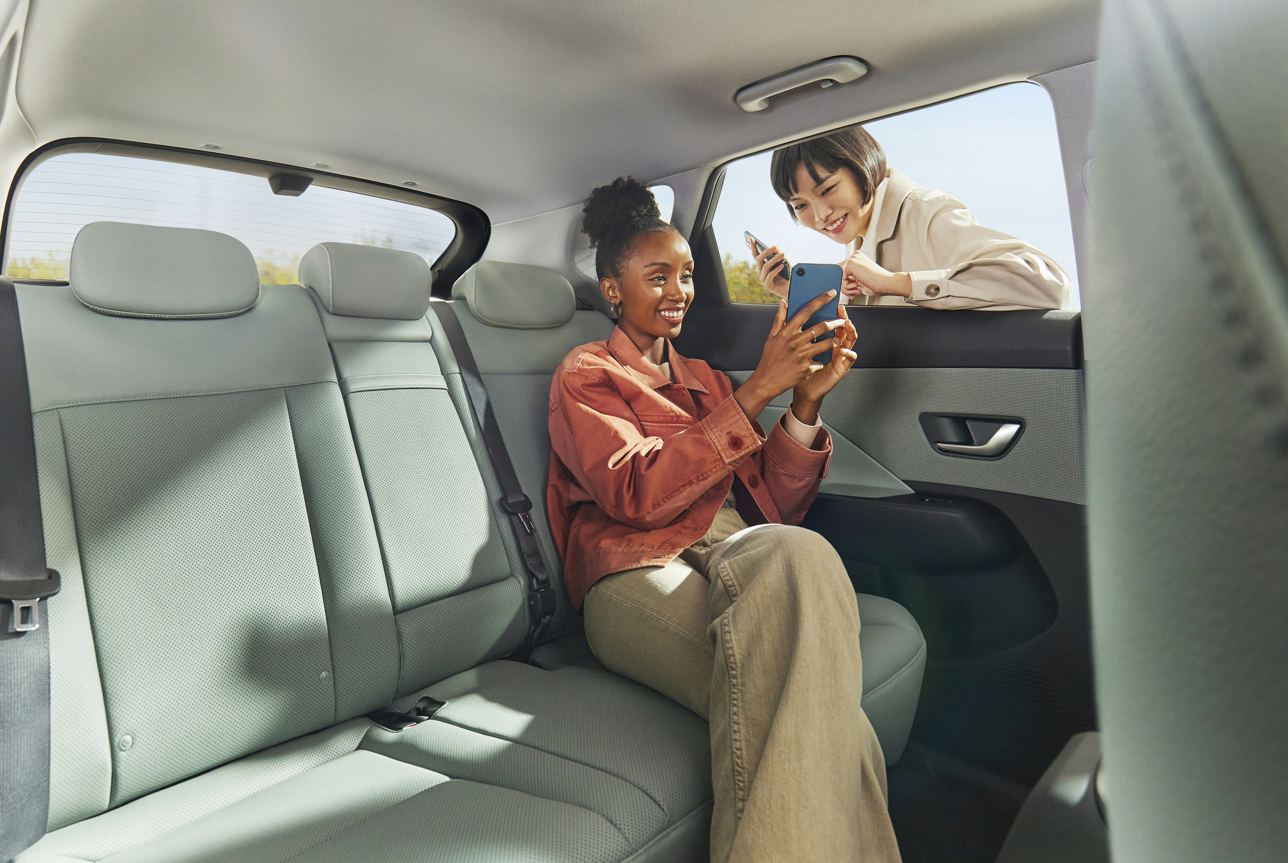 A woman sitting in the back seat of the Hyundai KONA showing her smartphone to a friend outside. 