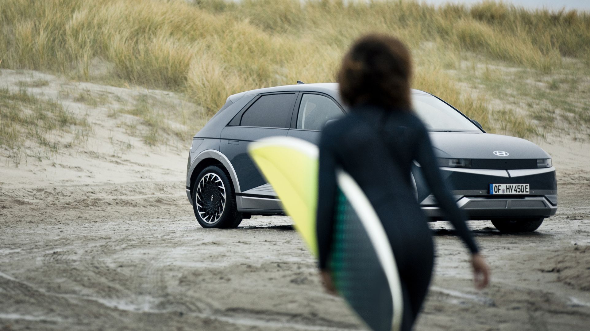 A surfer approaching her Hyundai IONIQ 5 electric vehicle parked on the beach.