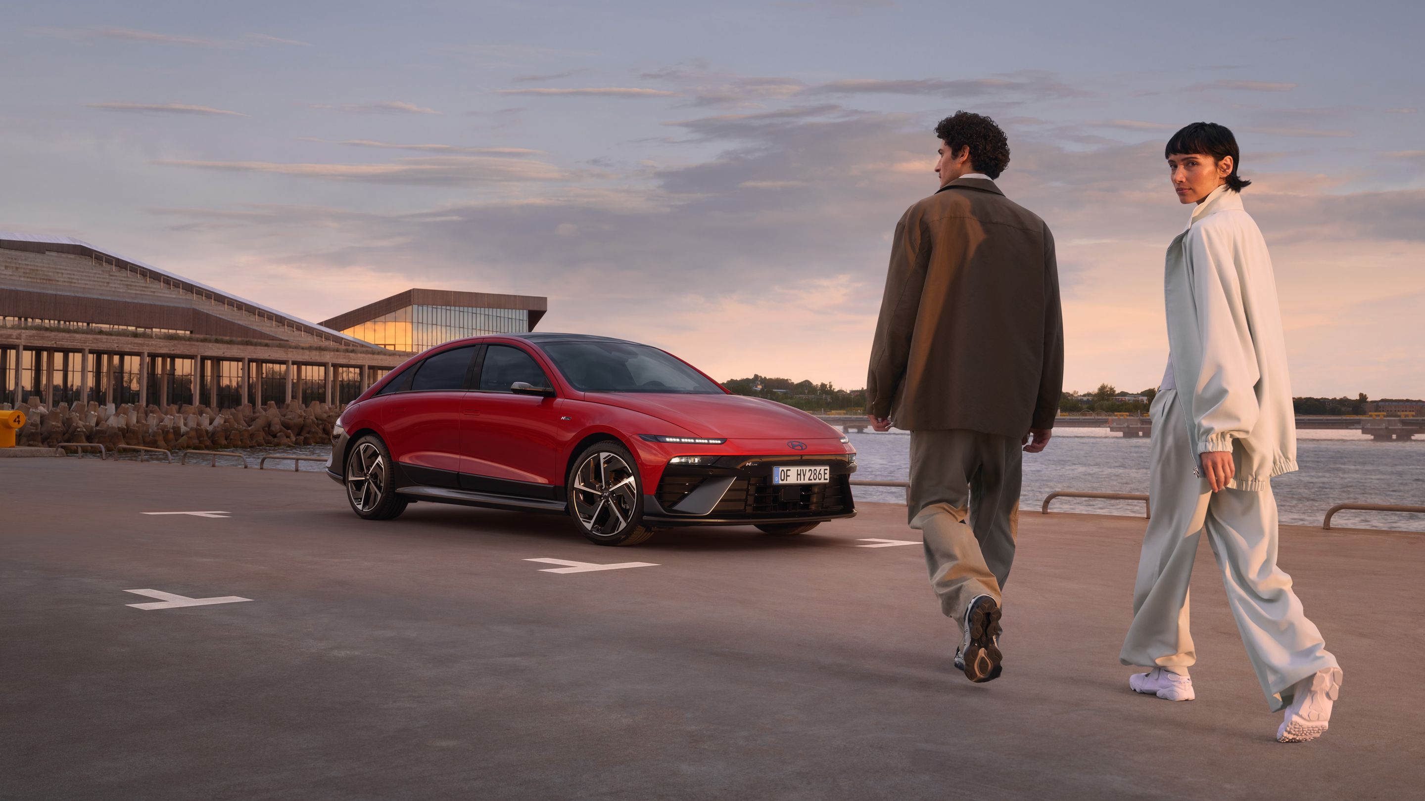 A man and a woman walking past a red Hyundai IONIQ 6 N Line parked next to the ocean.