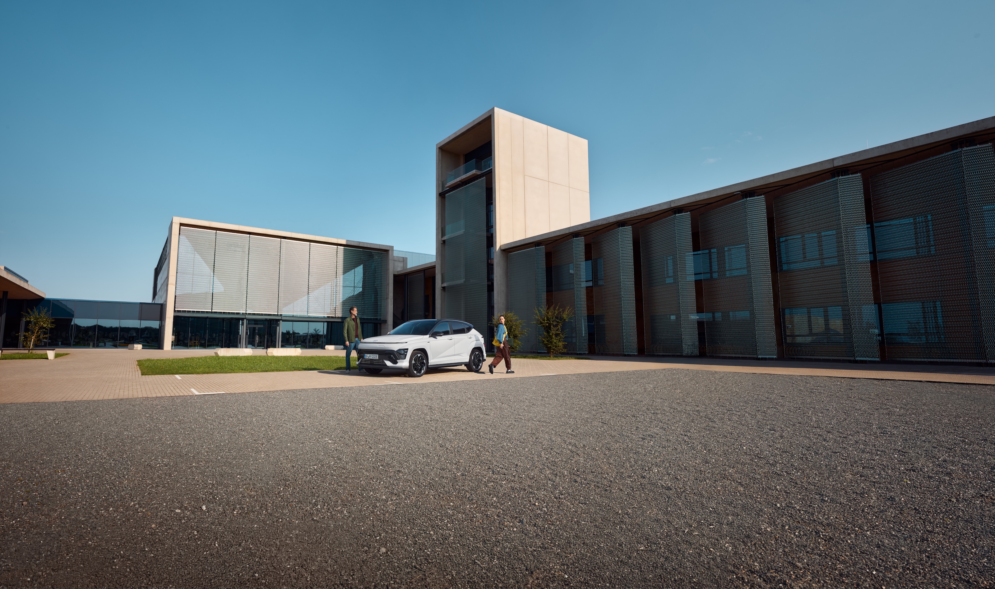 A white Hyundai Kona Electric car is parked with a man standing on the left side and a woman walking past on the right side. The background features a modern building with large windows.
