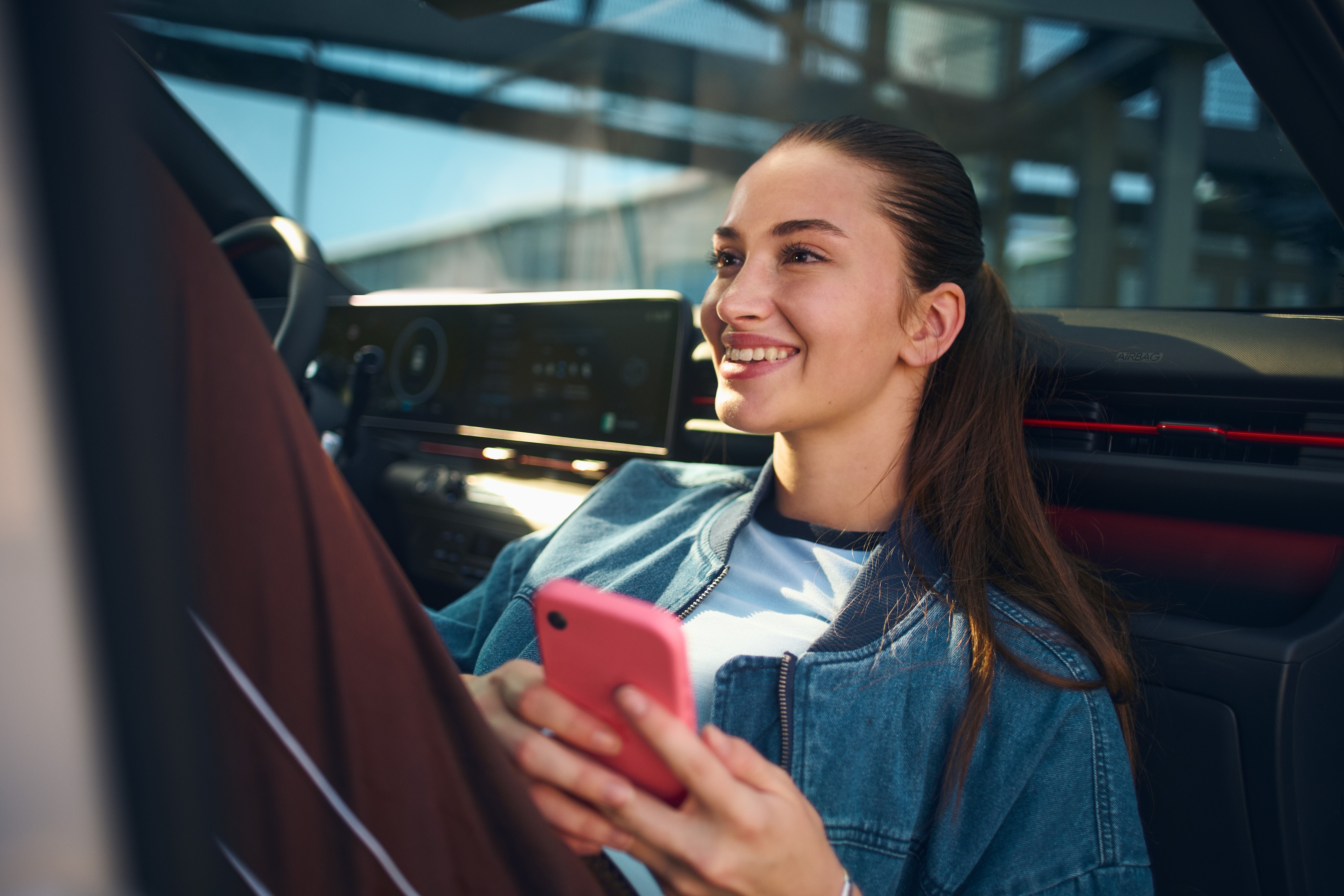 Person sitting in a car holding smartphone. The car dashboard with a large screen and controls is visible in the background.