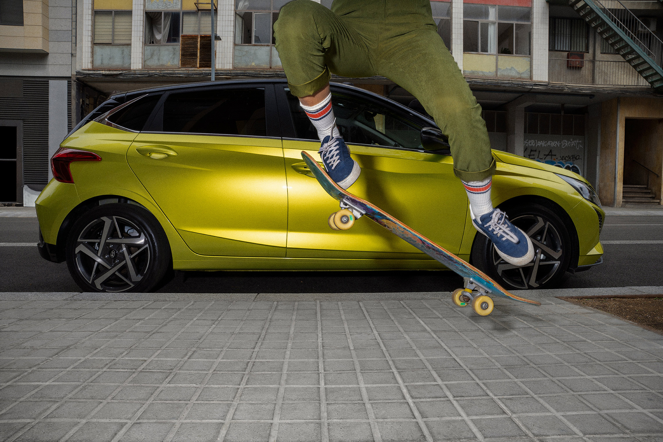 A person performing a skateboard jump with a Hyundai i20 in the background.	