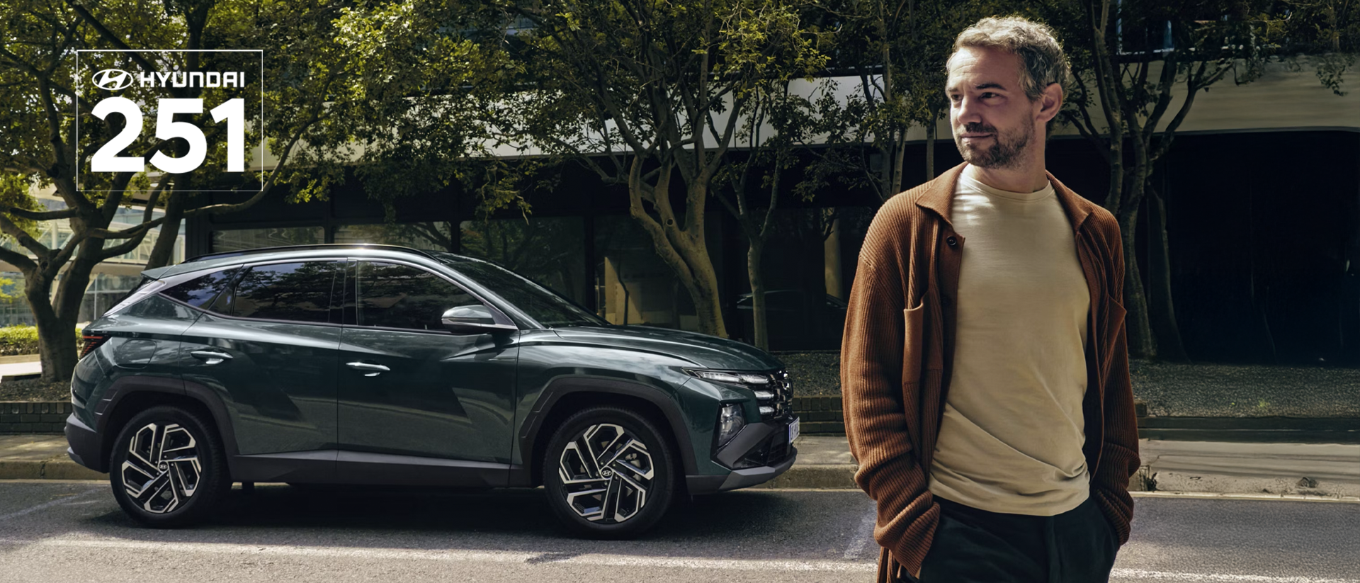 A man walking near a Hyundai TUCSON Hybrid parked by some trees.