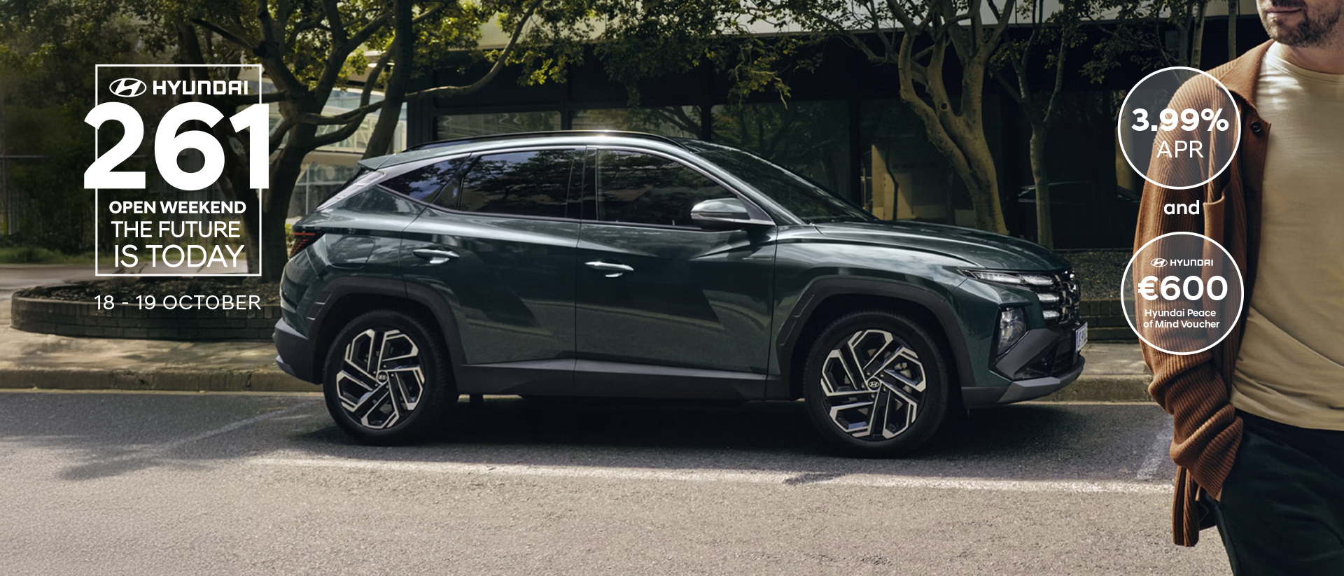 A man walking near a Hyundai TUCSON Hybrid parked by some trees.
